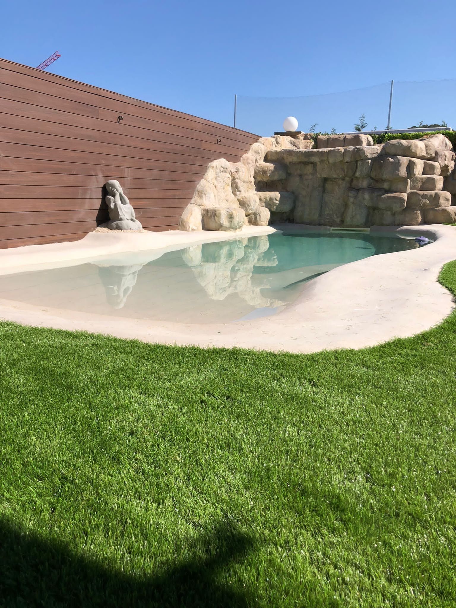 Pequeña piscina de arena rodeada de césped, pared alta al lado izquierdo, estructura de grandes piedras blancas al fondo