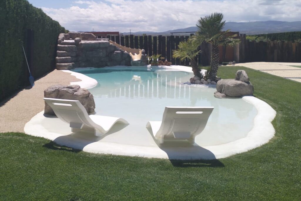 Piscina pequeña de arena rodeada de césped con dos tumbonas blancas a la entrada, seto alto a la izquierda, al fondo una estructura de piedra con escalera y un tobogán, a la mitad de la piscina en el lado derecho dos palmeritas con piedras alrededor. Pueden observarse las vistas en un día nublado
