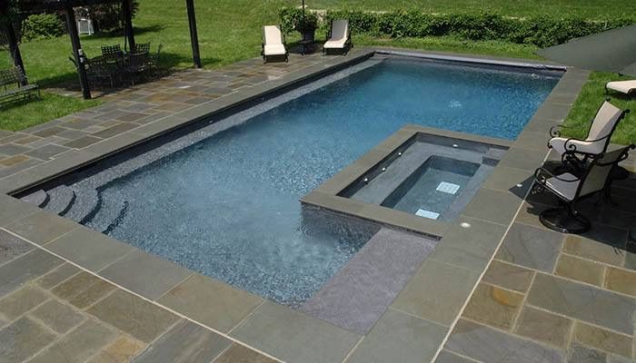 Piscina rectangular sobre suelo, con jacuzzi en un lateral, rodeada de plaqueta gris oscura, escalera de obra en la esquina izquierda, al fondo jardín con árboles con tumbonas y sillas en las que descansar