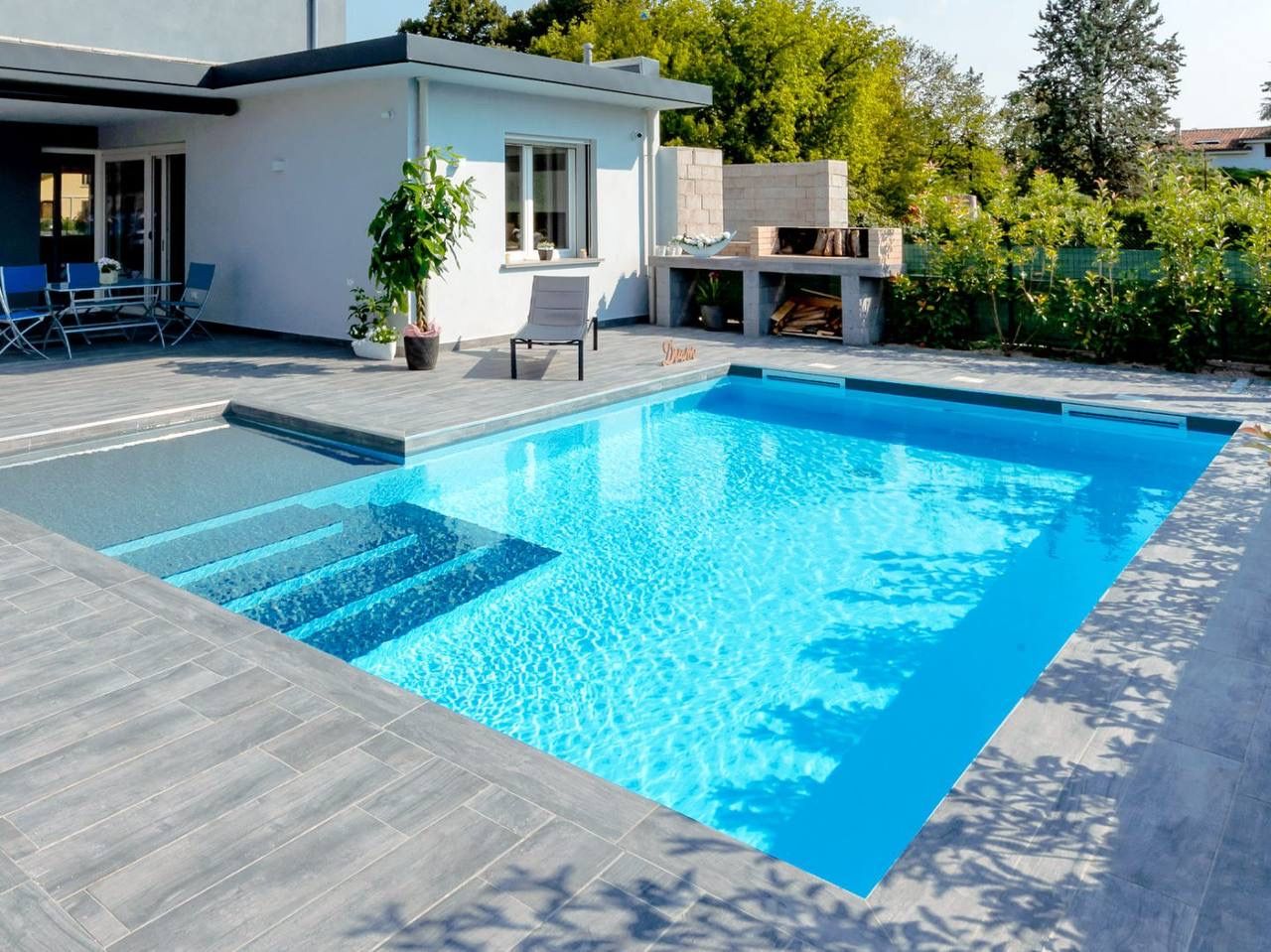 Piscina sobre suelo de tarima gris clara, con una amplia escalera, en el patio de una casa con porche en el que hay mesa y sillas para comer, al lado de la piscina hay una tumbona y varias plantas y al fondo vegetación