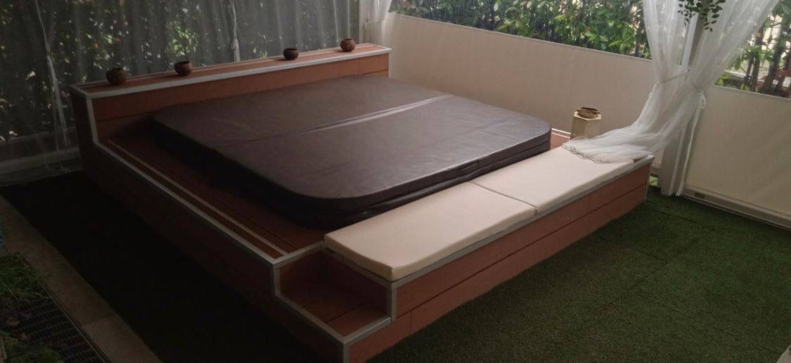 Jacuzzi cuadrado empotrado al aire libre, con tapa marrón, un banco a un lado y al otro especie de estante, con cortinas transparentes, rodeado de arbustos
