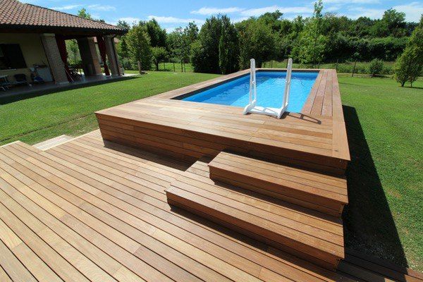 Piscina elevada de tarima color madera clara, sobre césped de un jardín con unos árboles de fondo.