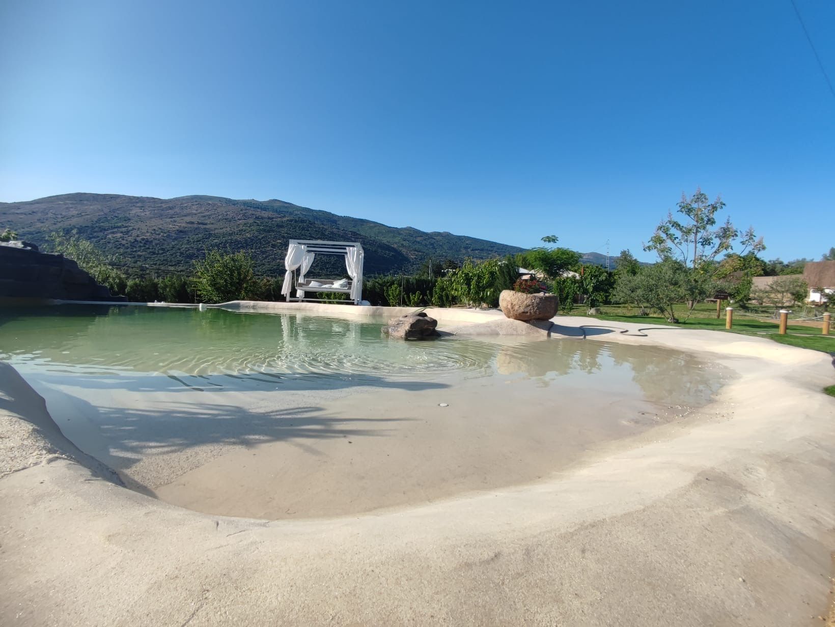 Gran piscina de arena blanca, rodeada de montañas, césped y árboles, para entrar a la misma hay un pequeño montículo de arena. Al fondo de la imagen cerca del agua hay una estructura con cortinas y un sofá blanco donde descansar